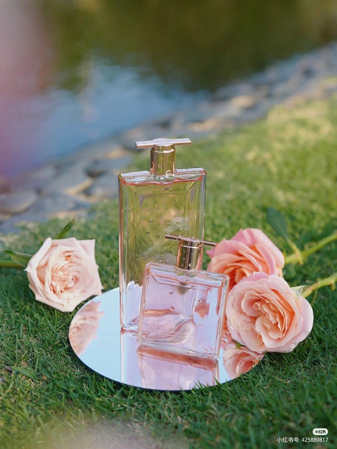 Two perfumes side by side one taller of the other on the grass close to a lake with 3 pink roses around those bottle. The background is blury. Photo credit: xxmhxx6 from Pinterest